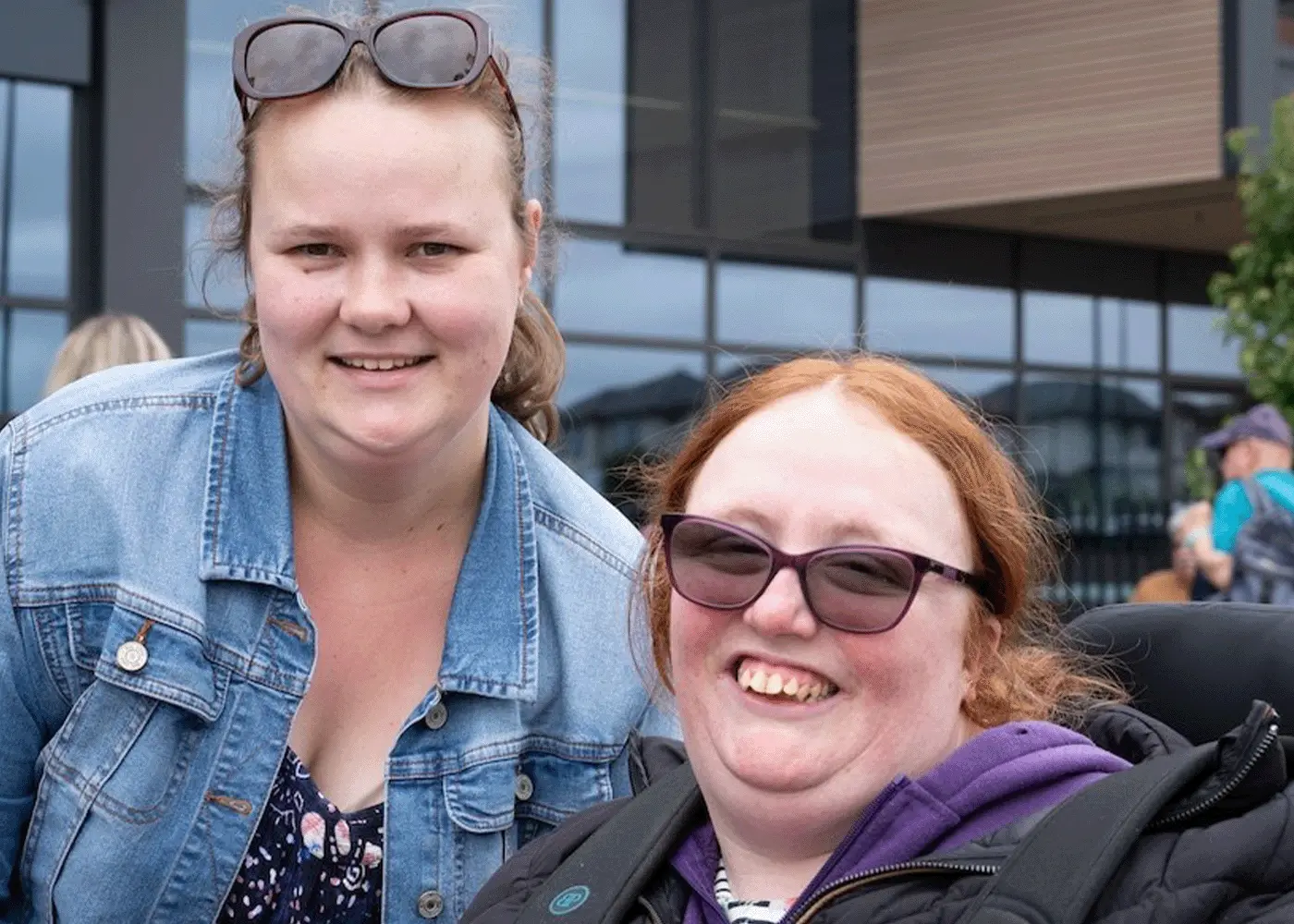 Two women, one in a wheelchair, smile at camera while at an outdoor event