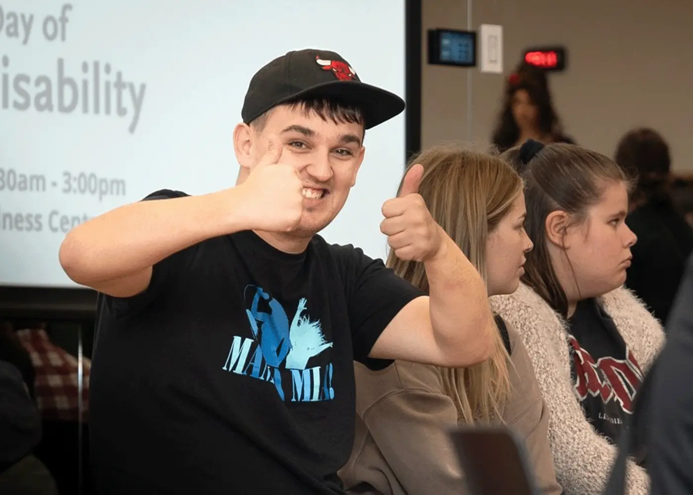 A young man wearing a baseball cap give the thumbs up sign while sitting in a meeting
